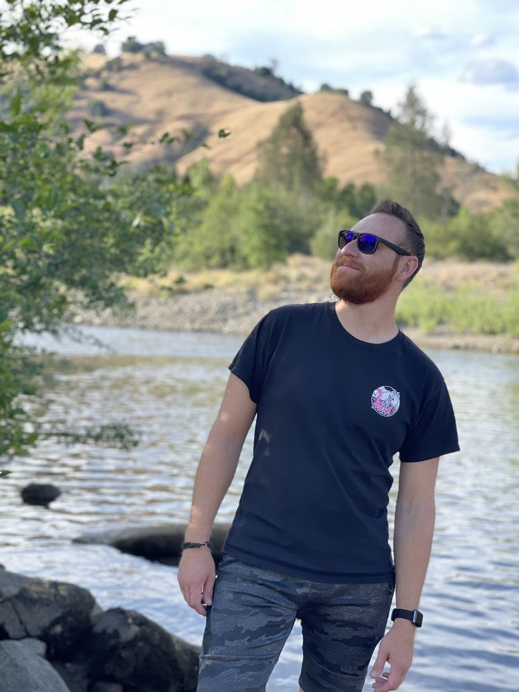 A man with a reddish beard and sunglasses stands by a body of water, looking up and to his right. He is wearing a black t-shirt with a small graphic on the chest, camouflage shorts, a watch, and bracelets. In the background, there are rolling hills, green trees, and a river. The weather appears sunny with some clouds in the sky.