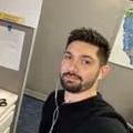 A selfie of a man with short brown hair and a beard, wearing a black shirt and white wired earbuds. He is looking directly at the camera in what appears to be an office cubicle, with a bulletin board and a water cooler map visible in the background.