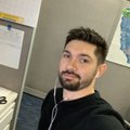 A selfie of a man with short brown hair and a beard, wearing a black shirt and white wired earbuds. He is looking directly at the camera in what appears to be an office cubicle, with a bulletin board and a water cooler map visible in the background.