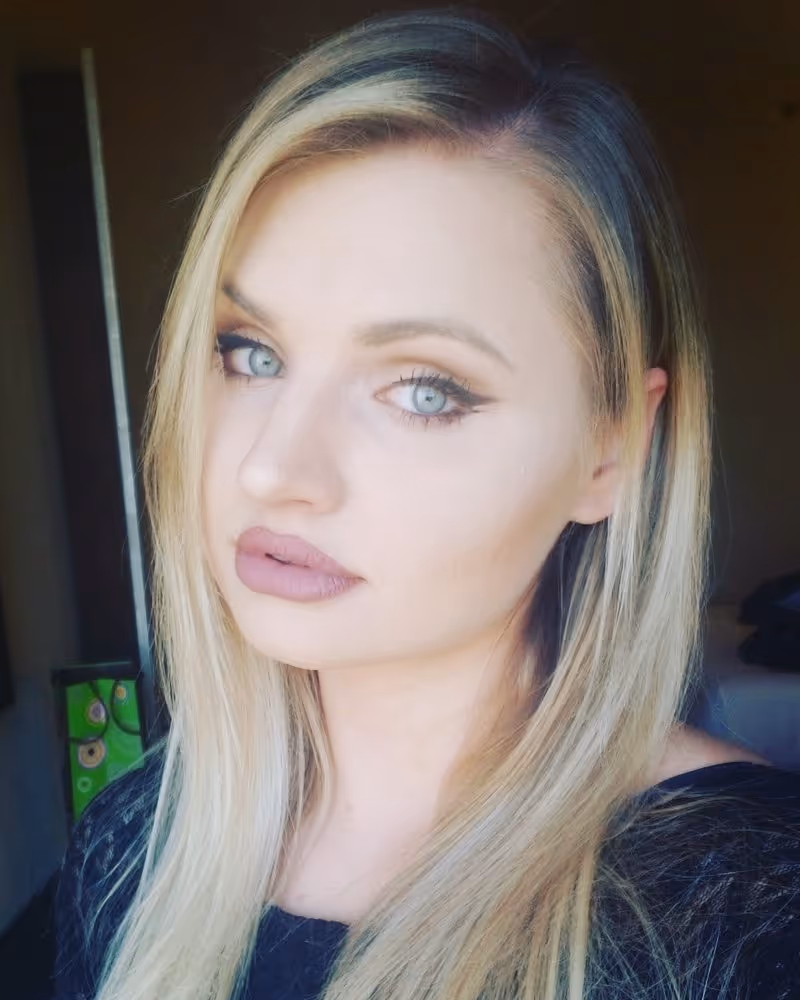 A selfie-style headshot of a woman with long, straight blonde hair and a dark top. She is wearing makeup, including dark eyeliner and lipstick, and is looking slightly away from the camera with a neutral expression. The lighting is soft, possibly from a window, and the background is dark and out of focus. 