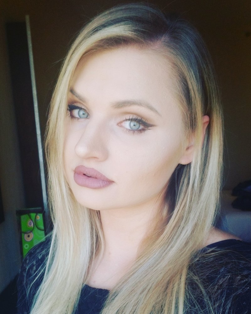A selfie-style headshot of a woman with long, straight blonde hair and a dark top. She is wearing makeup, including dark eyeliner and lipstick, and is looking slightly away from the camera with a neutral expression. The lighting is soft, possibly from a window, and the background is dark and out of focus. 