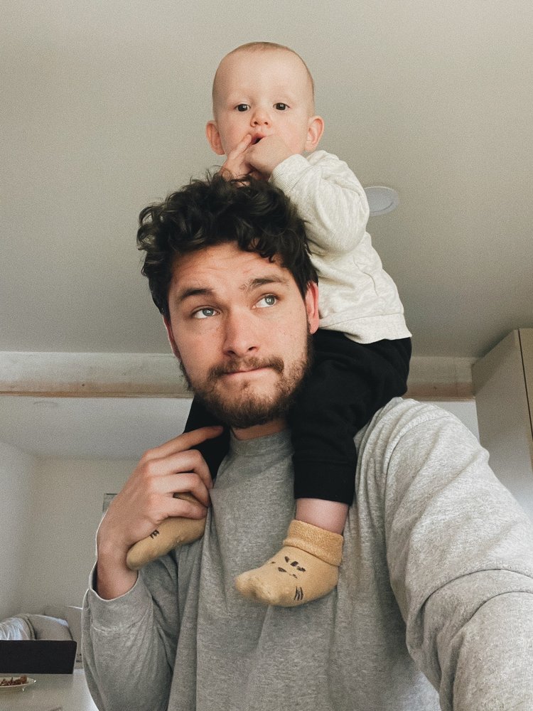 A man with dark, curly hair and a beard is giving a baby a piggyback ride on his shoulders. The baby, dressed in a light-colored top, is looking at the camera with a hand near its mouth.