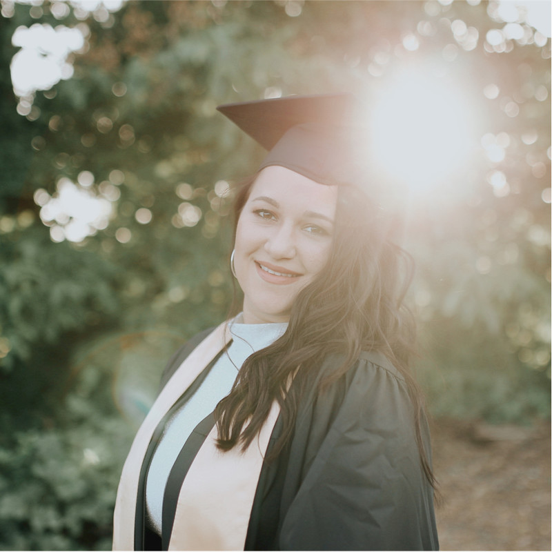 A graduate in a black cap and gown with a light stole smiles at the camera. The sun flares brightly behind her, creating a soft glow and backlighting her long hair. The background is blurred green foliage.