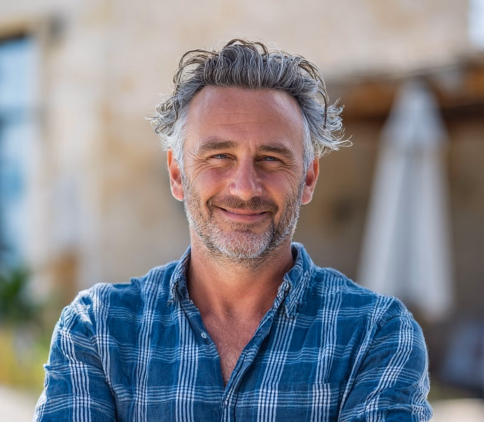 A man with a stylish haircut, featuring short sides and a swept-back top with light-colored or graying hair, smiles at the camera. He has light blue eyes and is wearing a denim button-up shirt.
