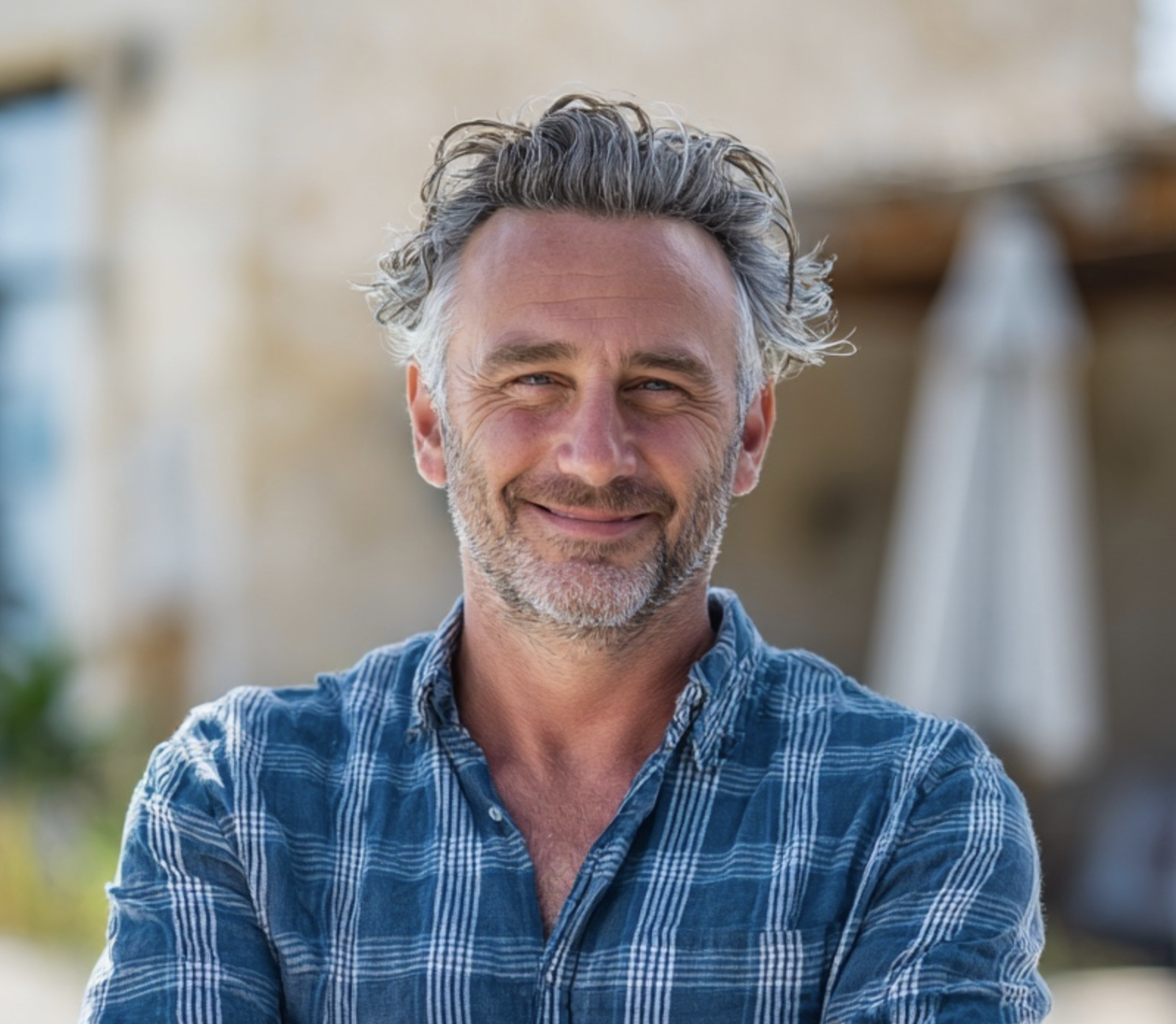 A man with a stylish haircut, featuring short sides and a swept-back top with light-colored or graying hair, smiles at the camera. He has light blue eyes and is wearing a denim button-up shirt.