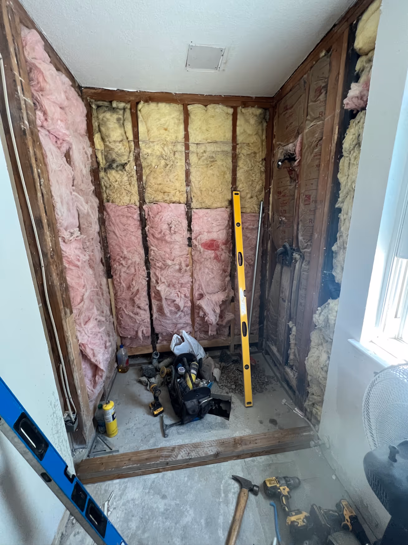 This image captures the interior of a bathroom or shower area mid-renovation, with the walls completely stripped down to the studs. The exposed wall cavity is partially filled with patches of both pink and yellow fiberglass insulation, revealing the bare structure of the room. A yellow spirit level stands upright against the back wall, suggesting that work on framing or leveling is underway. The floor is concrete or subfloor, covered in dust, with various tools, including drills, hammers, and construction materials, scattered around the work area. The raw state of the room, illuminated by natural light from a window on the right, clearly indicates that the space is being prepared for new walls and finishes.