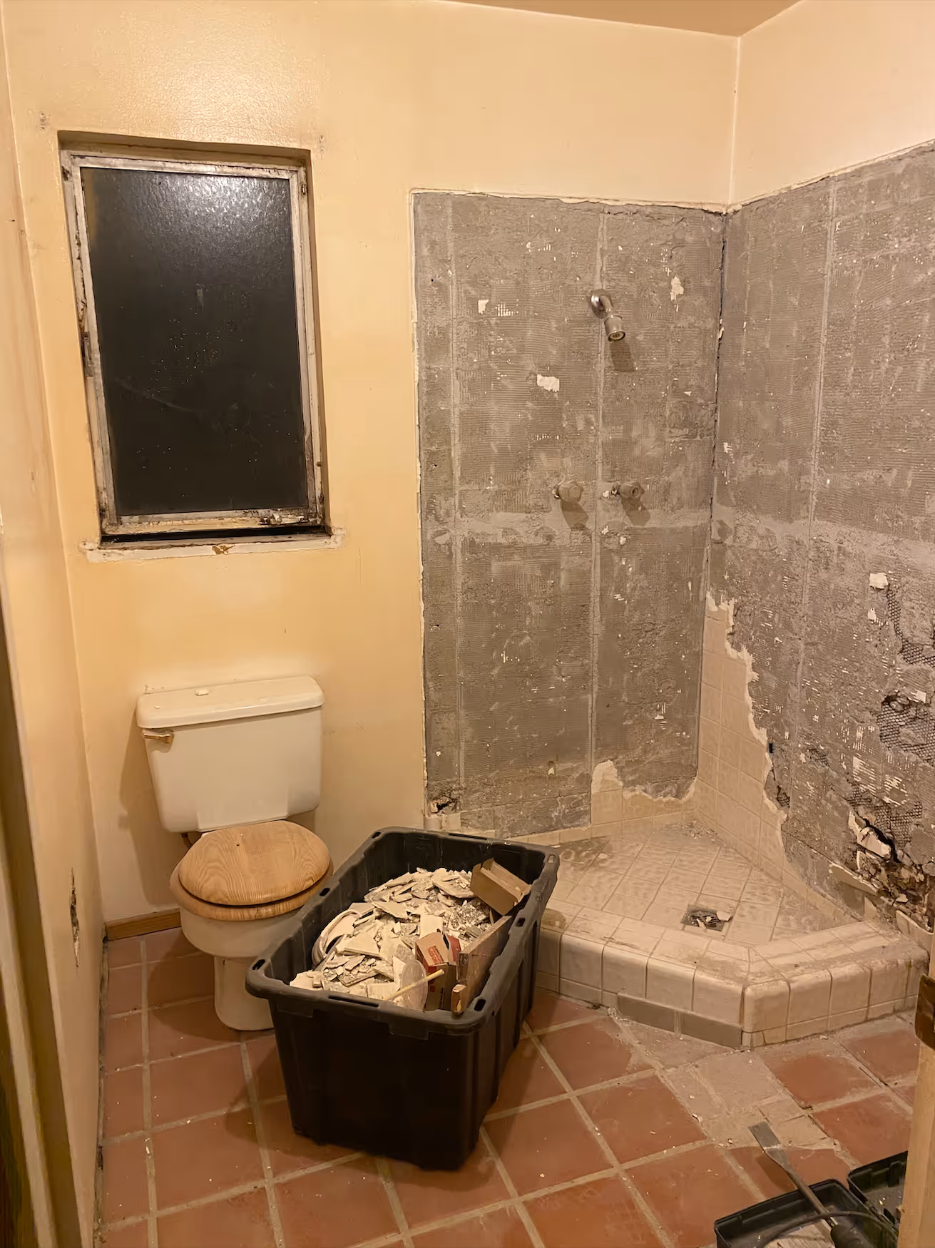 
This image shows a bathroom undergoing demolition and renovation, highlighting the "before" state of the space. The floor is covered in dusty, reddish-brown square tiles, and the walls are painted a dull yellow. The shower area is in the process of being stripped, with the original tiles removed to expose the cement board and lathe beneath. A white toilet with a wooden seat remains in the corner next to a small, dark window. In the center of the floor sits a large black tote bin filled with construction debris, including pieces of old tile and plaster, clearly signaling that the room is mid-project and preparing for a complete overhaul.