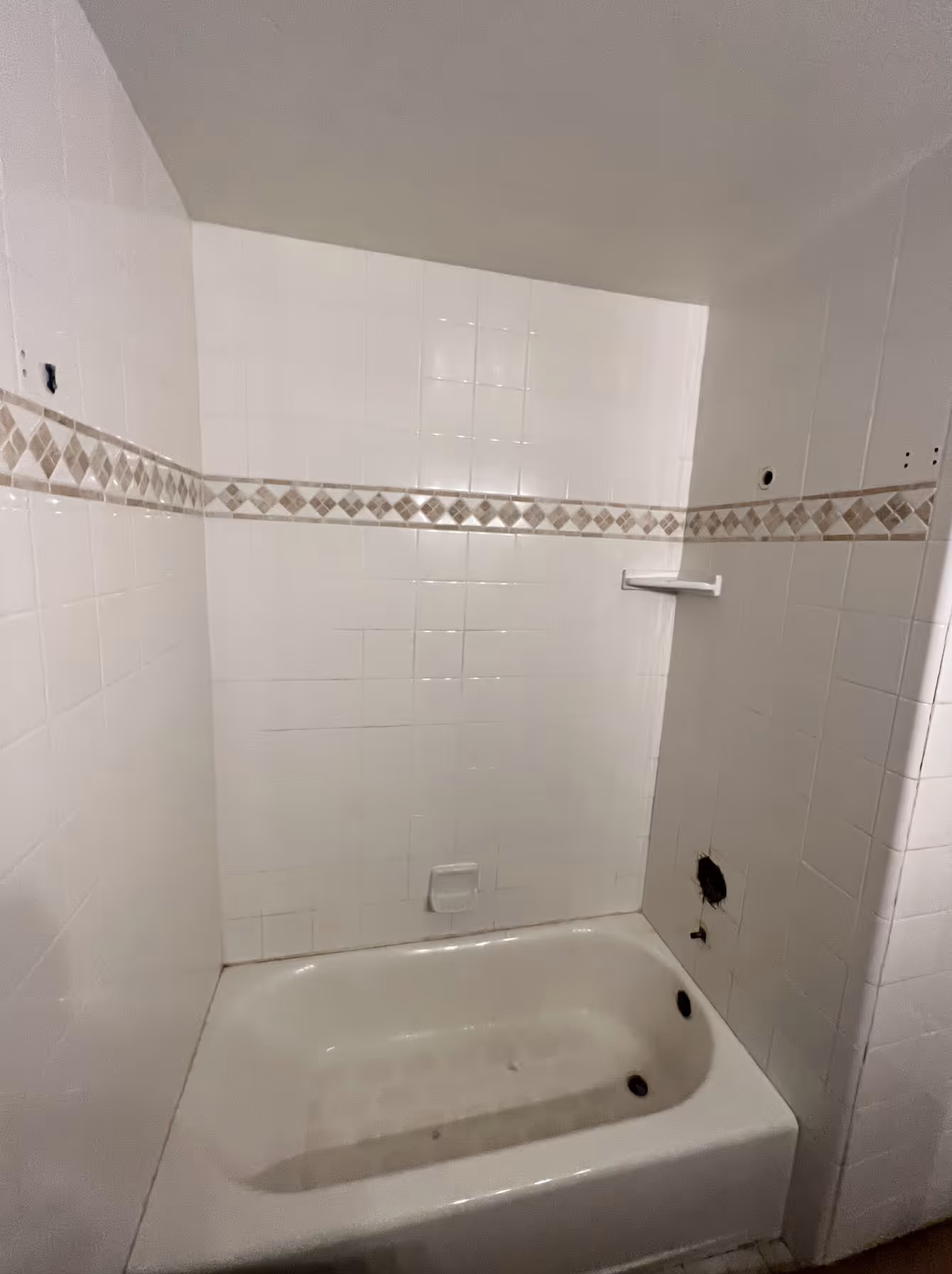 This image is a straight-on shot of an existing bathtub and shower enclosure, appearing to be an older style that is either waiting for renovation or has recently been stripped of fixtures. The shower area is completely tiled with square white ceramic tiles and features a decorative horizontal band of beige and brown diagonal accent tiles running around the perimeter. The white bathtub is visible at the bottom, and the tiled walls show evidence of removed accessories, with small holes indicating where a shower curtain rod and other fixtures were previously mounted. The scene has a slightly dated look, capturing the space before a modern update.