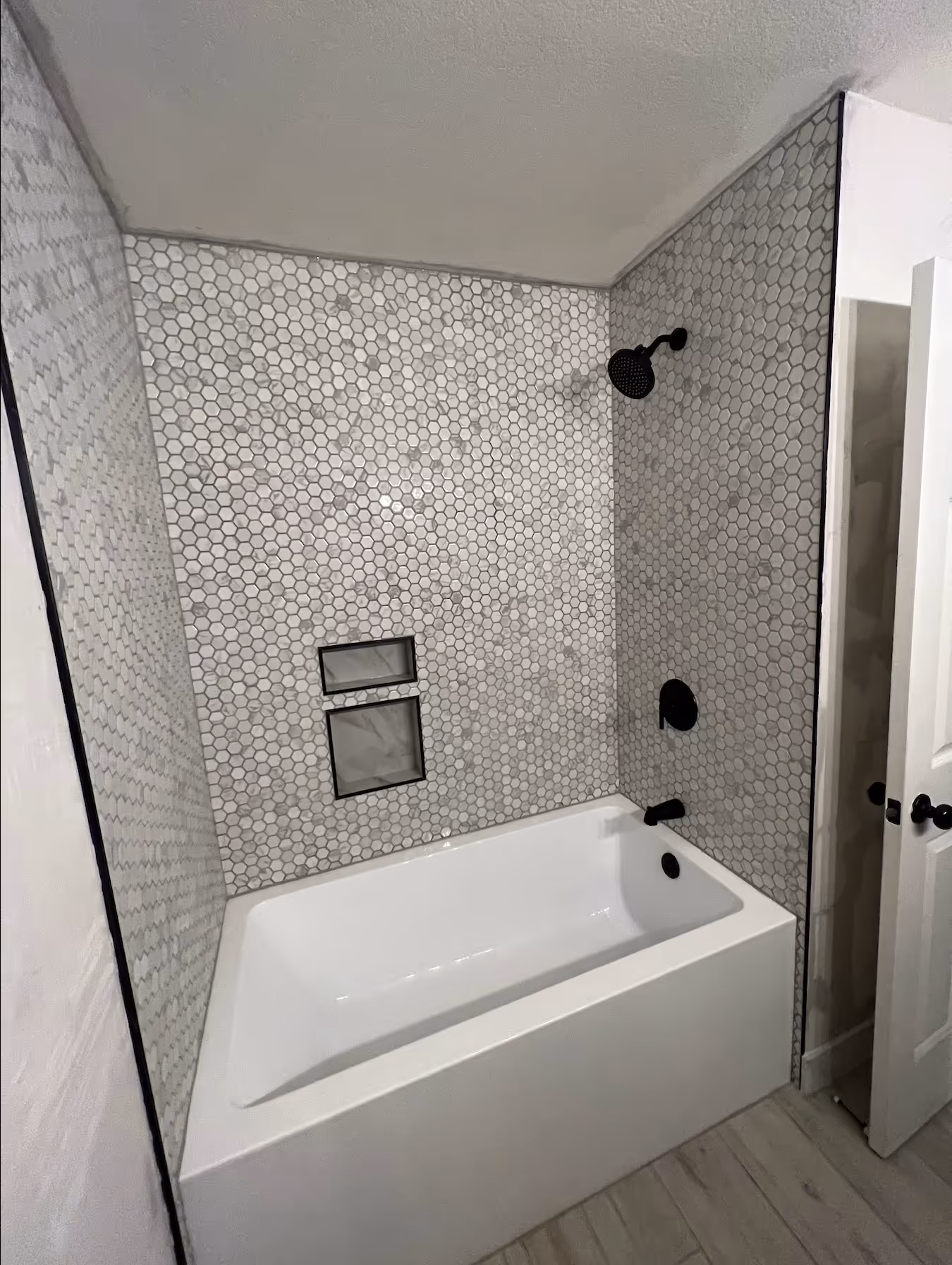 
This image showcases a newly renovated bathtub and shower enclosure with a striking, contemporary design. The walls are completely covered in small, white and light-gray hexagonal mosaic tiles (penny rounds), creating a textured and uniform pattern. A modern white bathtub is installed below, and the space features a double recessed niche in the back wall, framed in black and lined with a subtle marble-look tile for accent and storage. All the shower fixtures, including the showerhead, valve, and spout, are finished in matte black, providing a sharp, stylish contrast against the light-colored tiling. The overall aesthetic is clean, modern, and highly textural.
