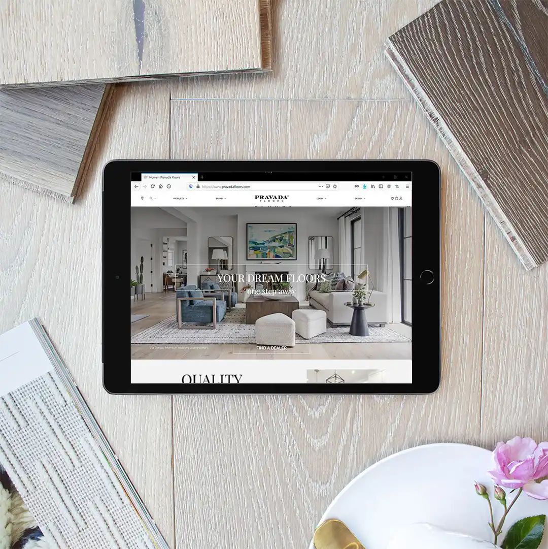 A tablet featuring the hero design of Pravada Floors website. The tablet lays on a wooden flooring surrounded by decorative wooden pieces and a decor magazine.