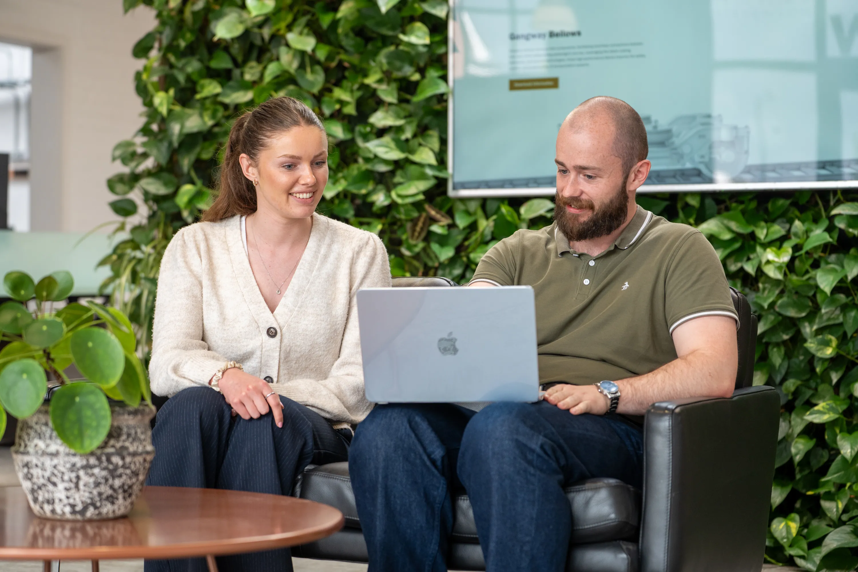 Smiling man and woman sitting on leather chairs looking at a laptop in a plant-filled office.