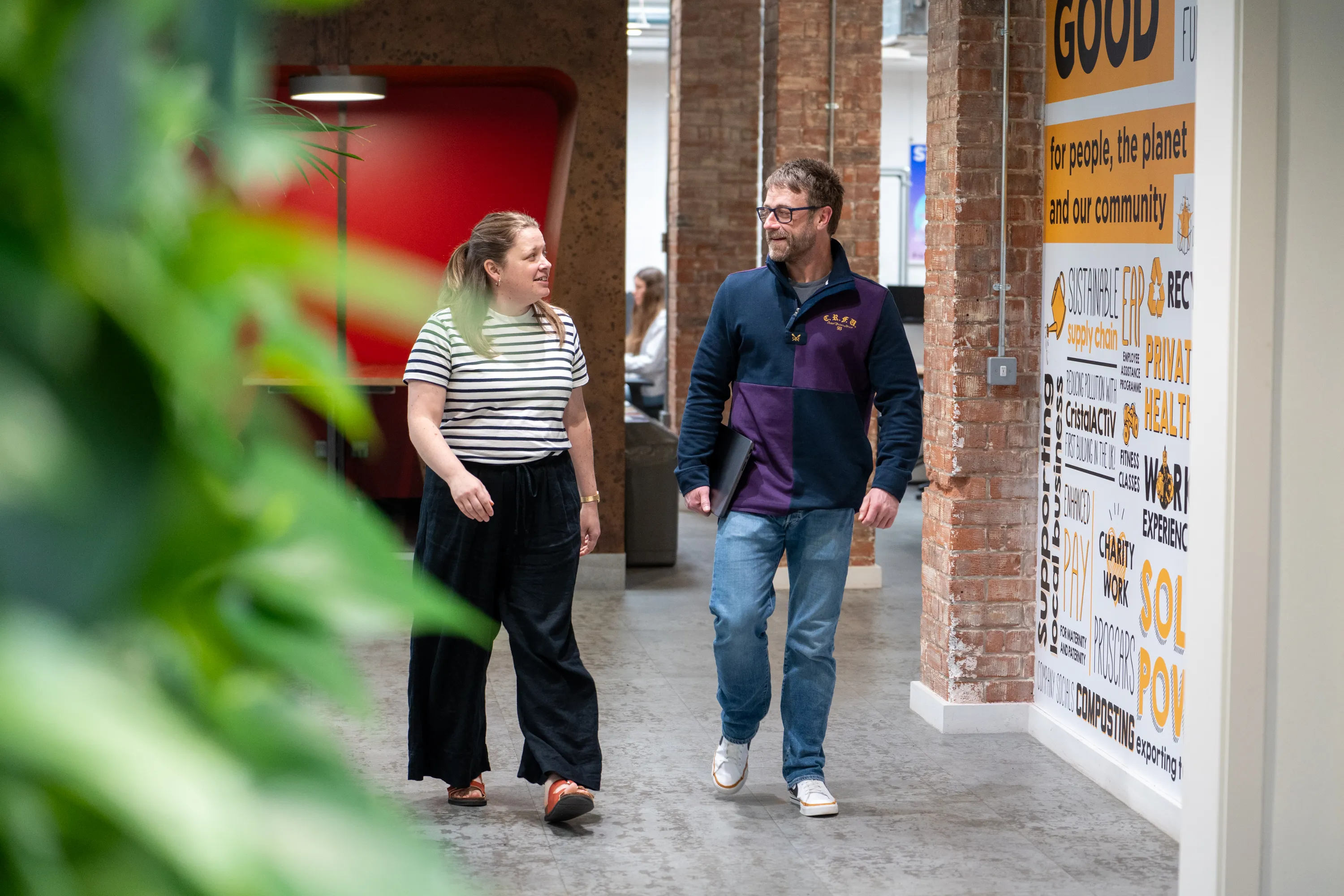 Two people walking and talking in a modern office hallway with brick walls and a large poster.