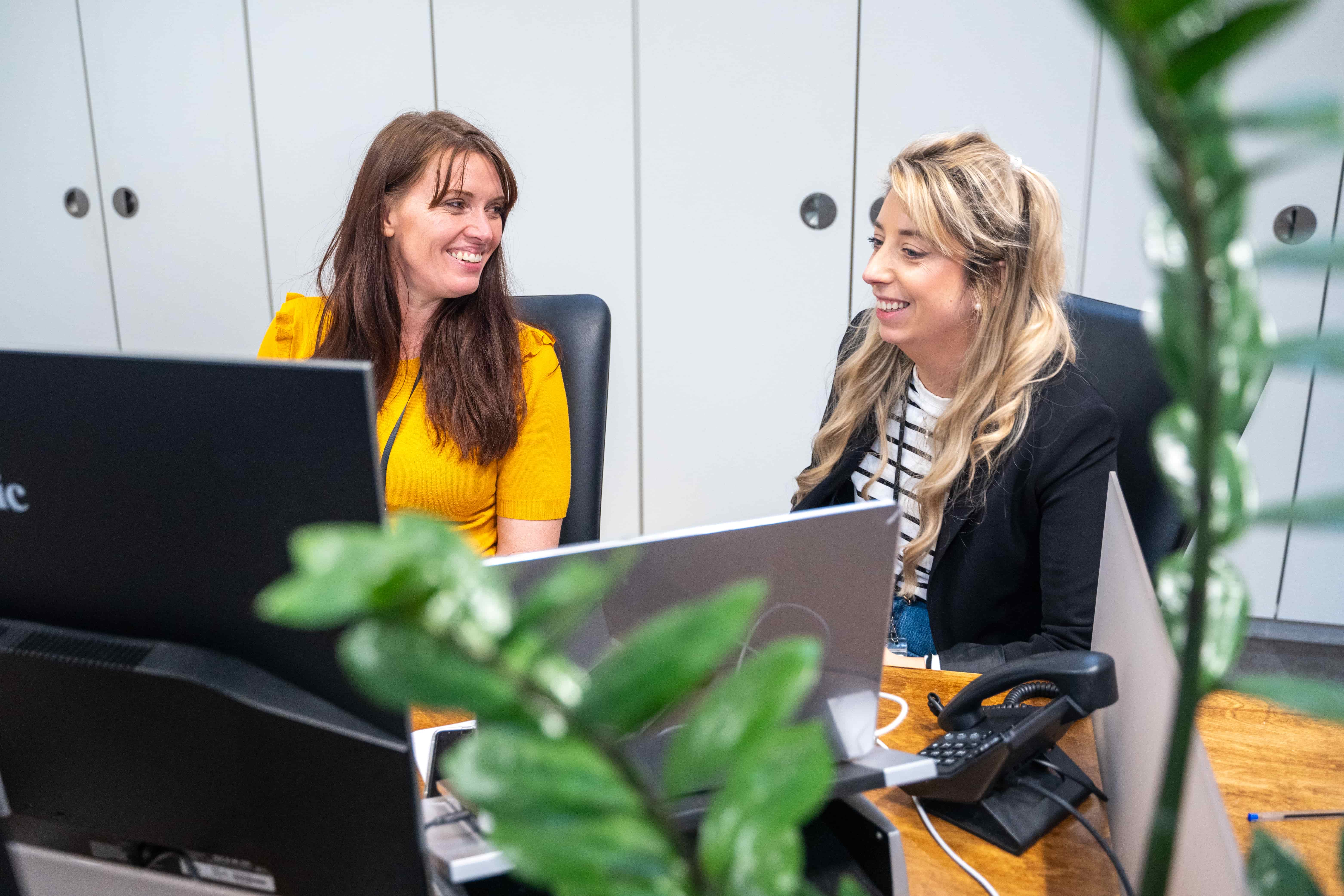 Two women sitting at desks with laptops, smiling and talking in a modern office setting.