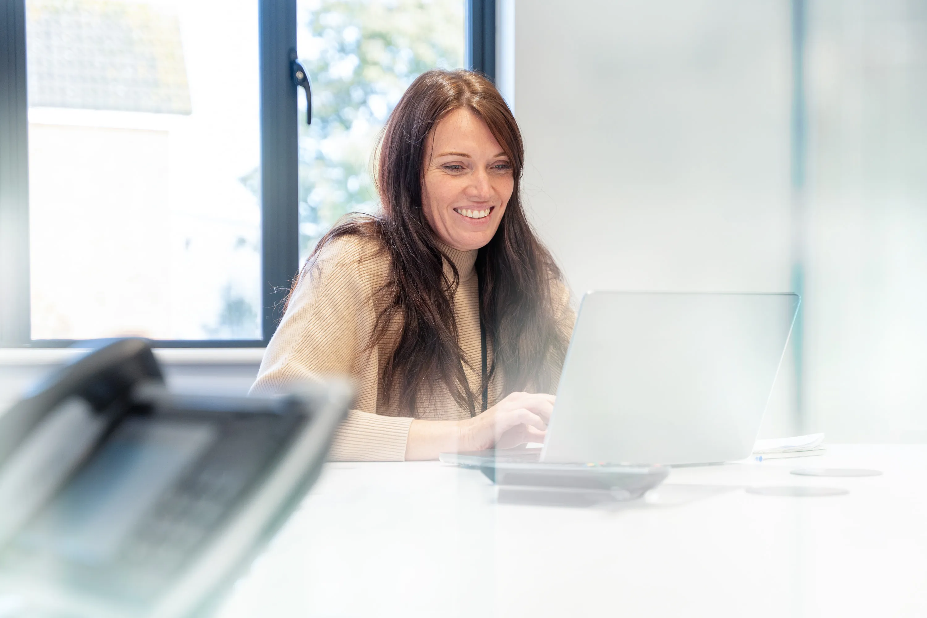 Smiling woman with long brown hair working on a laptop at a bright office desk near a window.