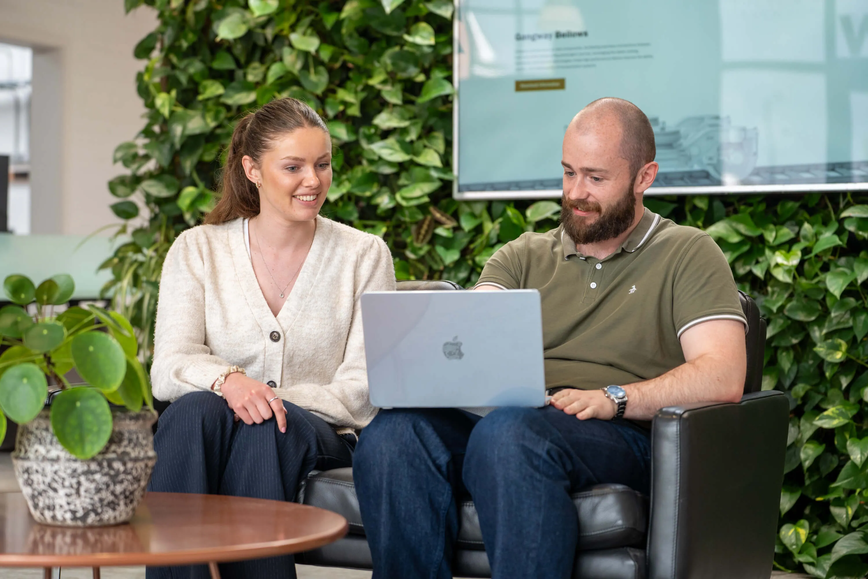 Smiling man and woman sitting on a black couch looking at a laptop with green plants in the background.