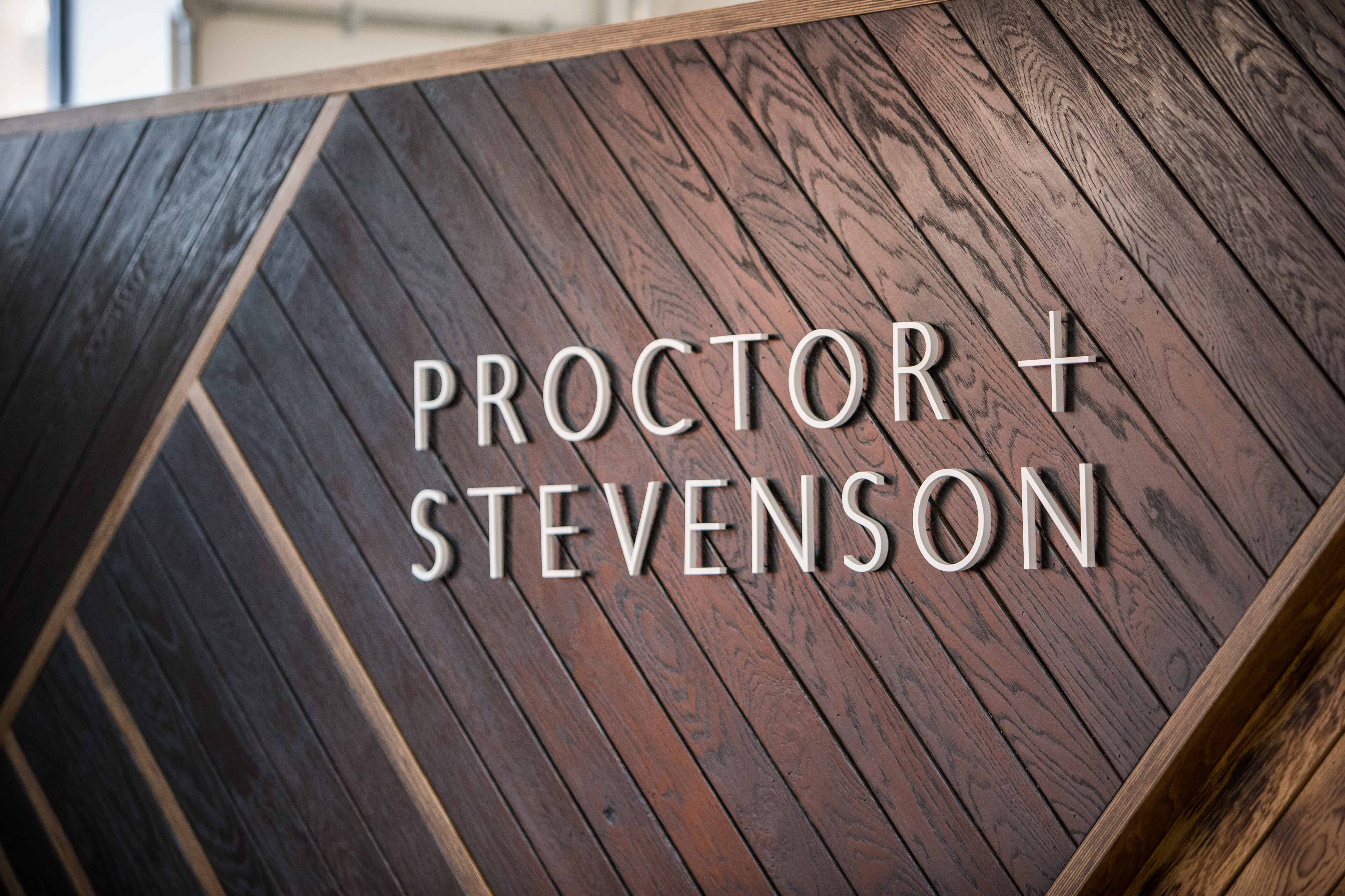 Wall with dark wooden panels arranged diagonally and raised white letters spelling PROCTOR + STEVENSON.