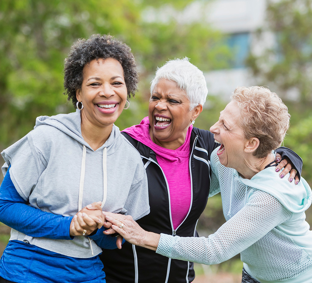 Three older women dressed in athletic clothing smiling and laughing outdoors while embracing each other.