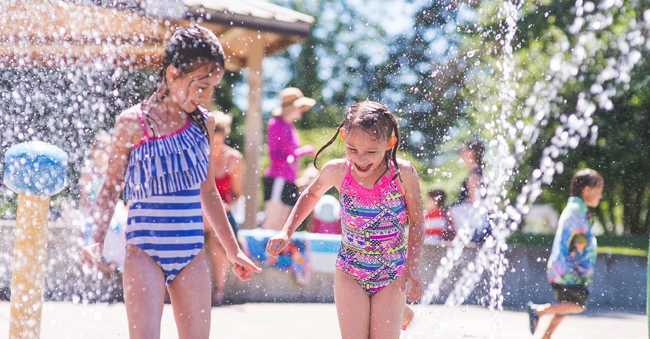 Two young girls in colorful swimsuits playing and laughing in a water splash park on a sunny day.