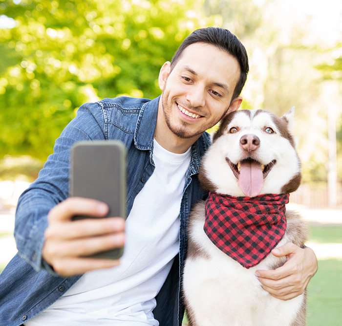 Man in a denim jacket taking a selfie with a happy Siberian Husky wearing a red checkered bandana in a sunny park.