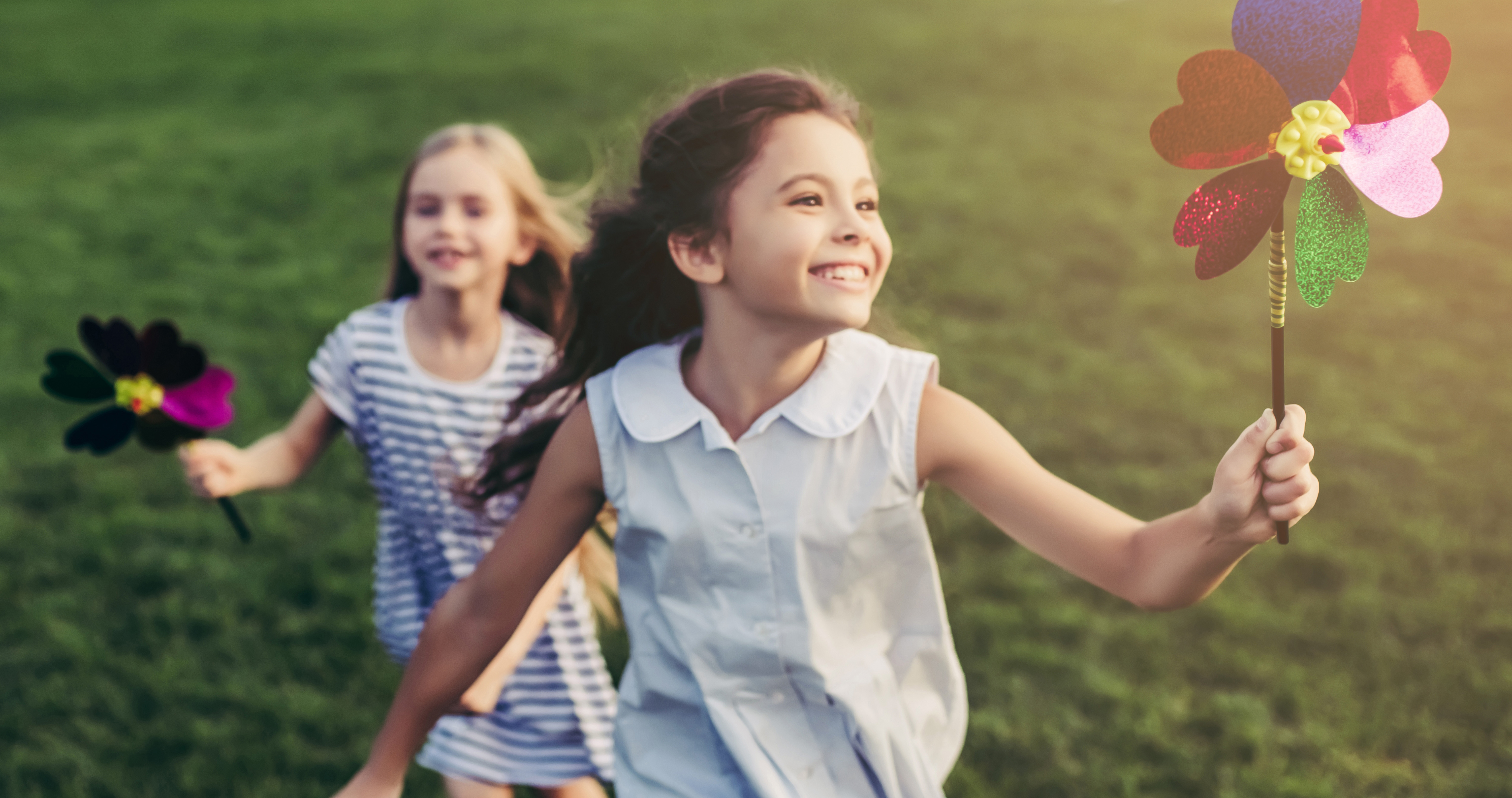 Two smiling girls running on grass holding colorful pinwheels on a sunny day.