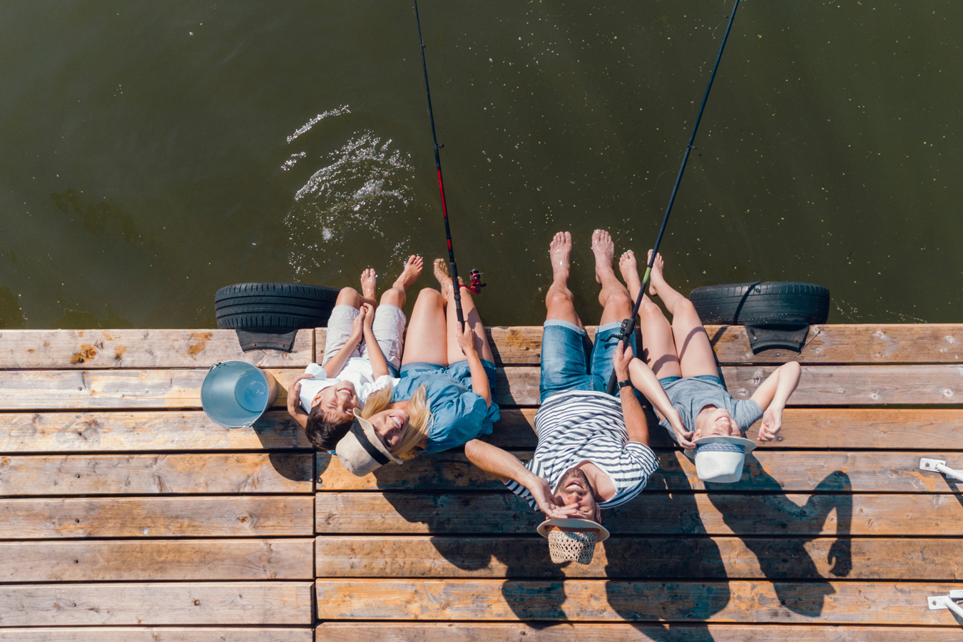Family of four sitting on a wooden dock fishing, with feet dangling above the water on a sunny day.
