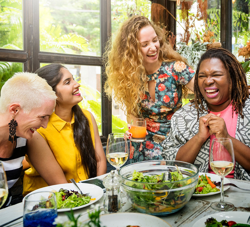 Four women laughing and enjoying a meal together at a table with salad bowls and drinks in a bright, plant-filled room.