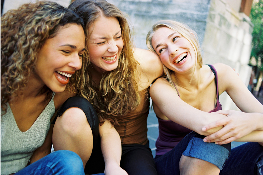 Three young women sitting close together outdoors, laughing joyfully.