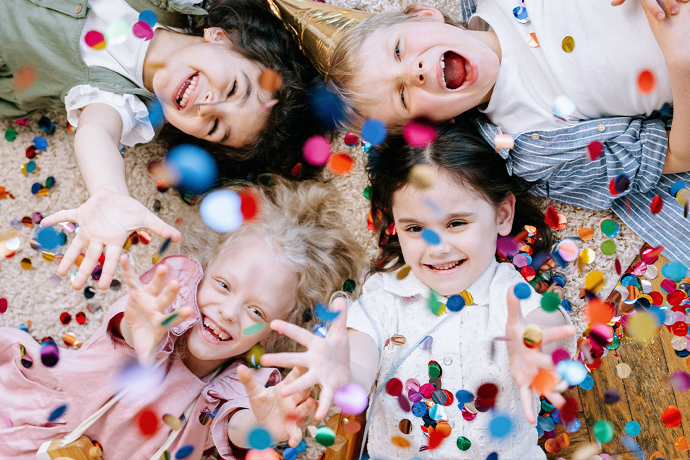 Four children lying on the floor smiling and reaching out as colorful confetti falls around them during a celebration.