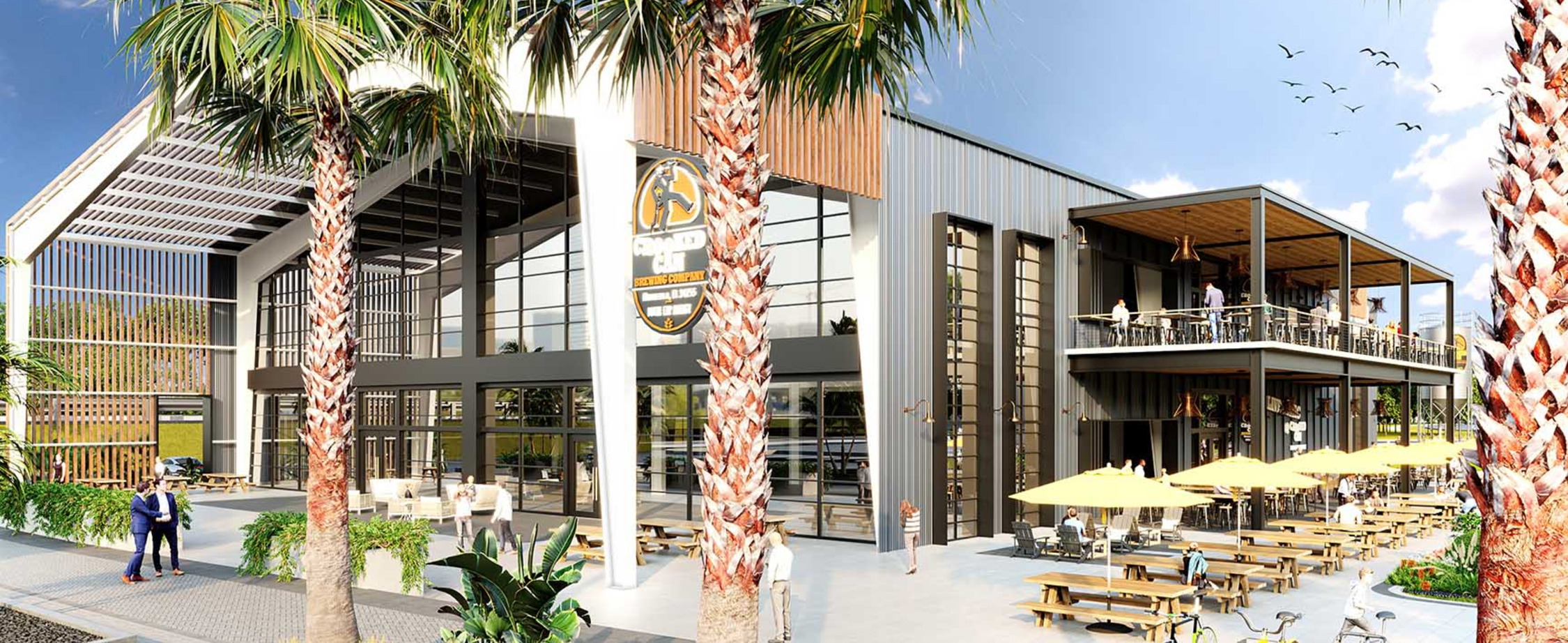 Modern building with large glass windows, outdoor seating areas with yellow umbrellas, picnic tables, palm trees, and people walking and sitting outside.