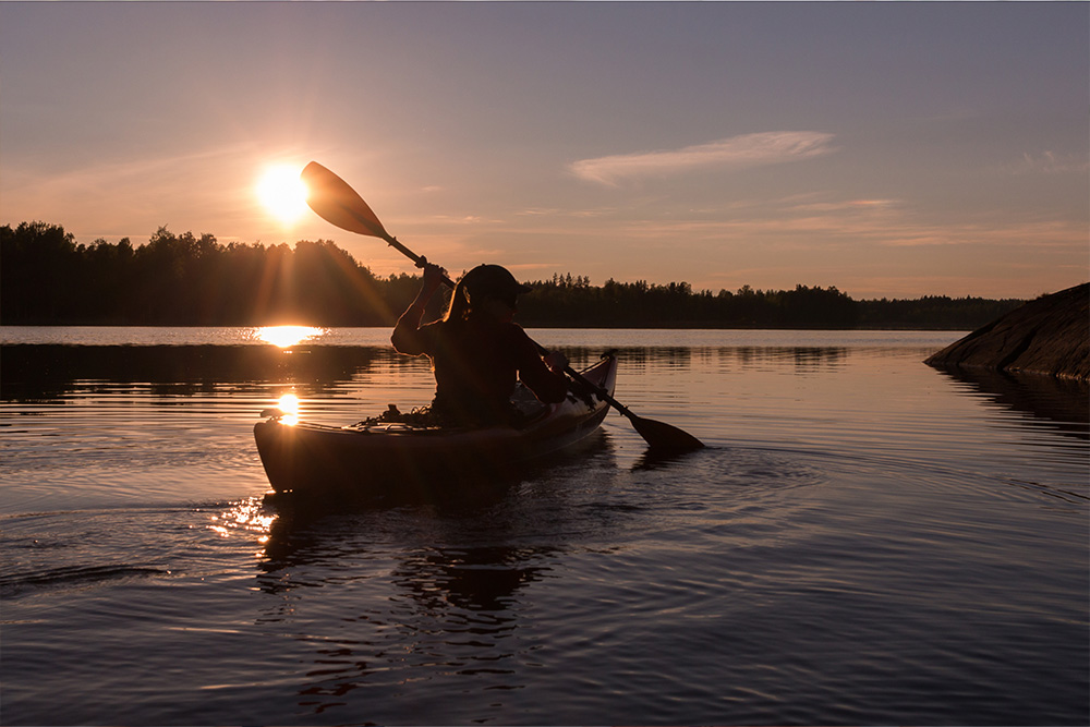 Person paddling a kayak on calm water at sunset with trees in the background.
