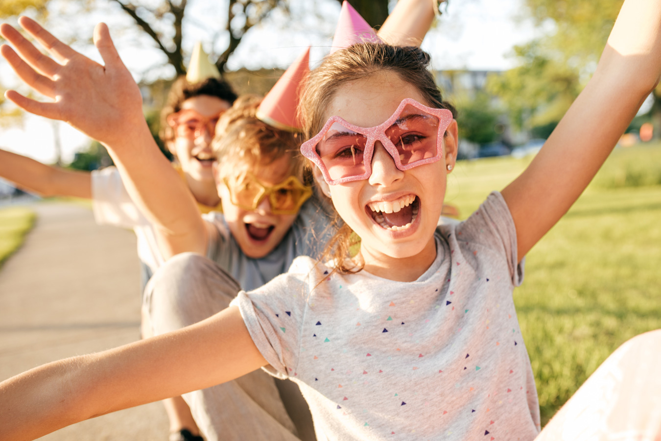 Three children wearing colorful party hats and oversized star-shaped sunglasses, smiling and raising their arms outdoors on a sunny day.