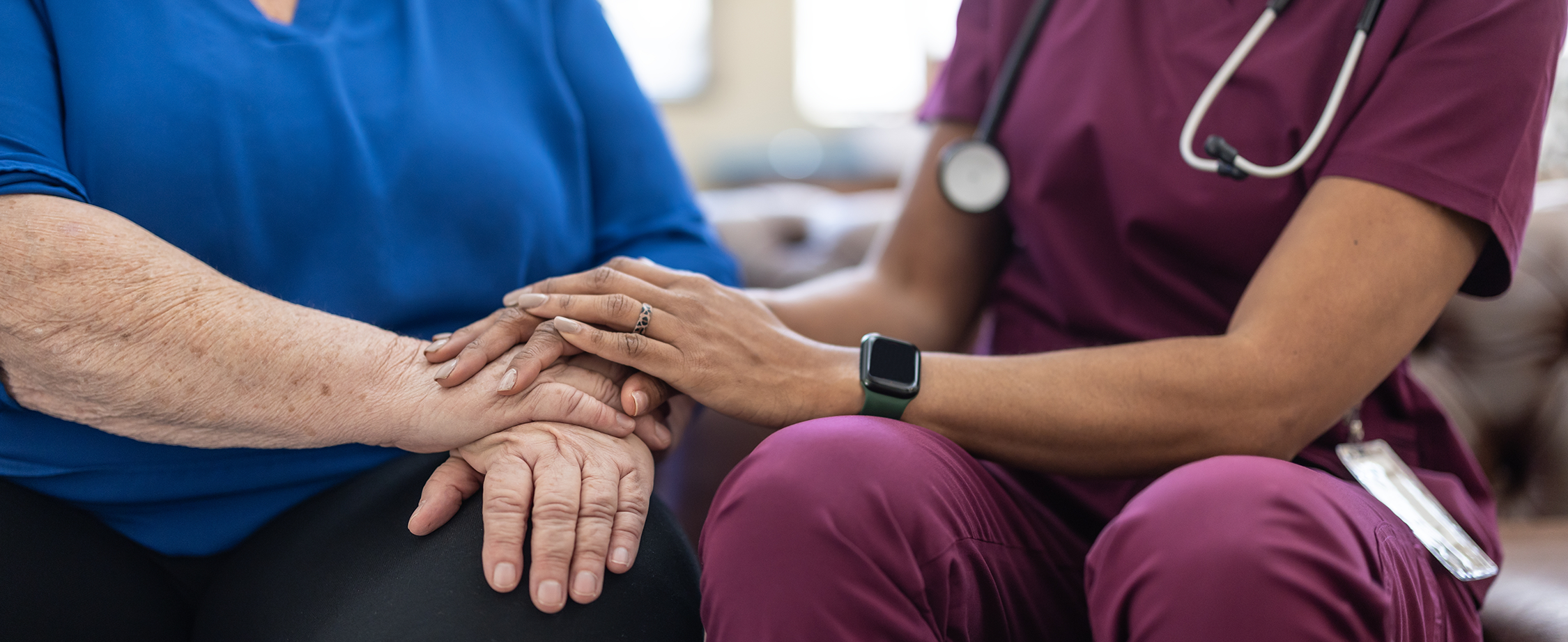 Healthcare professional in maroon scrubs holding the hands of an elderly person wearing a blue shirt in a comforting gesture.