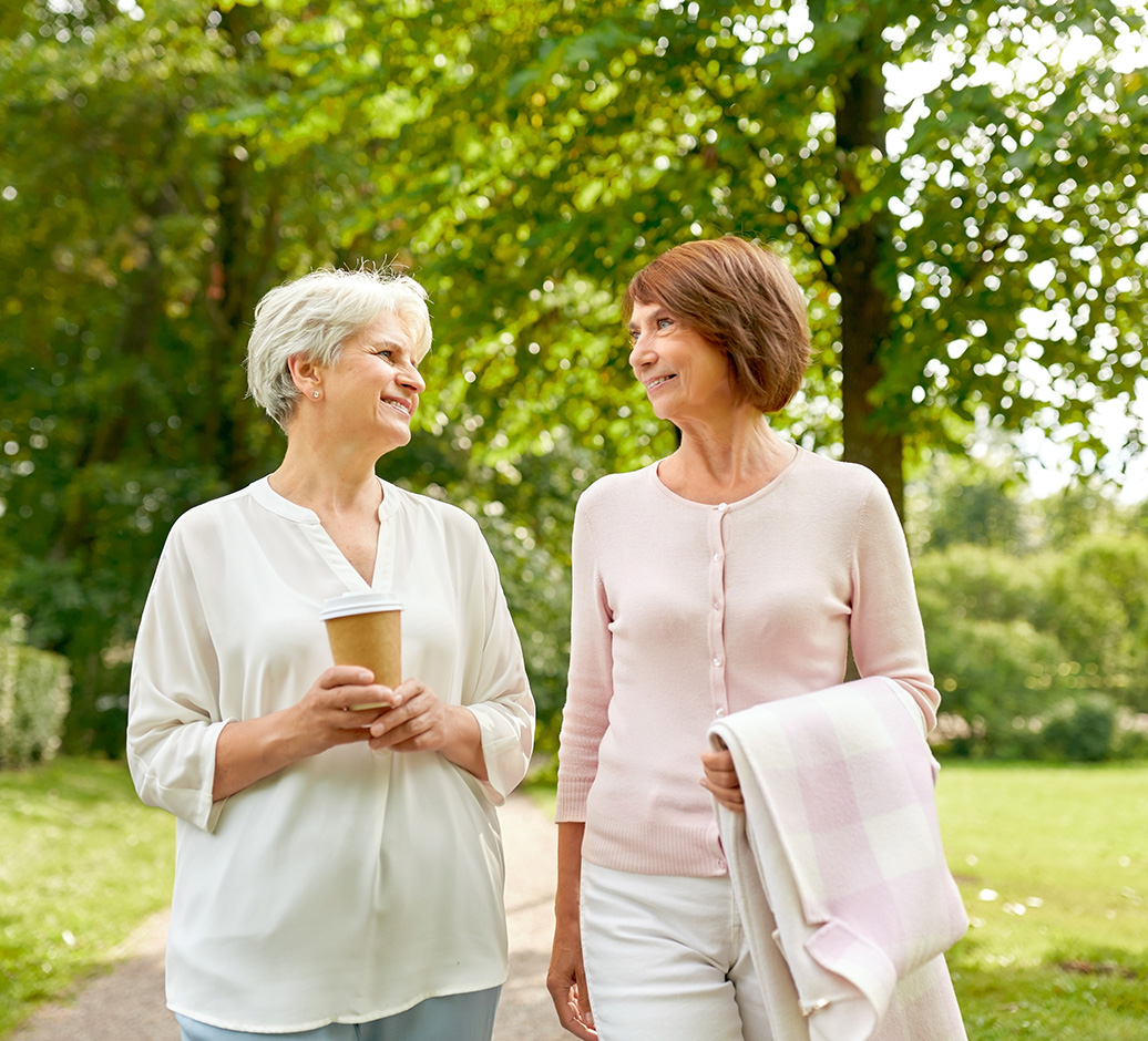 Two mature women smiling and walking in a green park, one holding a coffee cup and the other carrying a folded blanket.