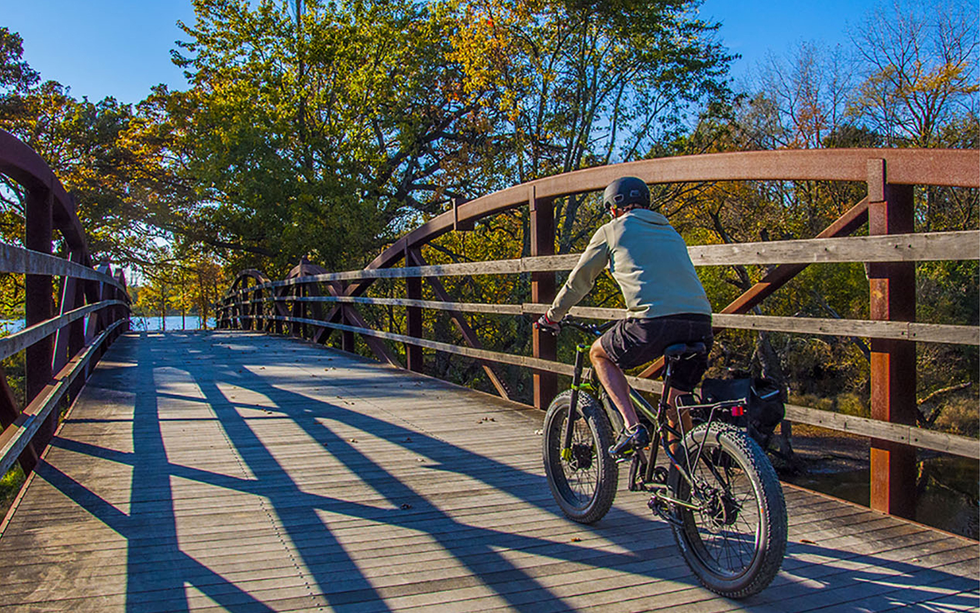 Person wearing a helmet riding a fat-tire bike across a wooden bridge surrounded by autumn trees under a clear blue sky.