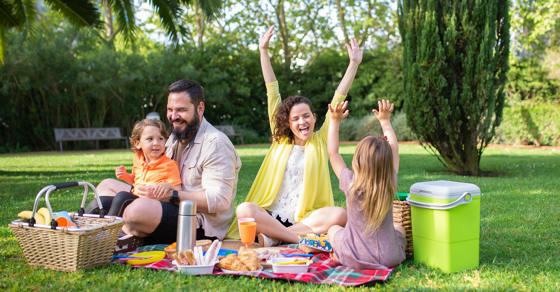 Family of four having a picnic on a red plaid blanket in a green park with food and a picnic basket.