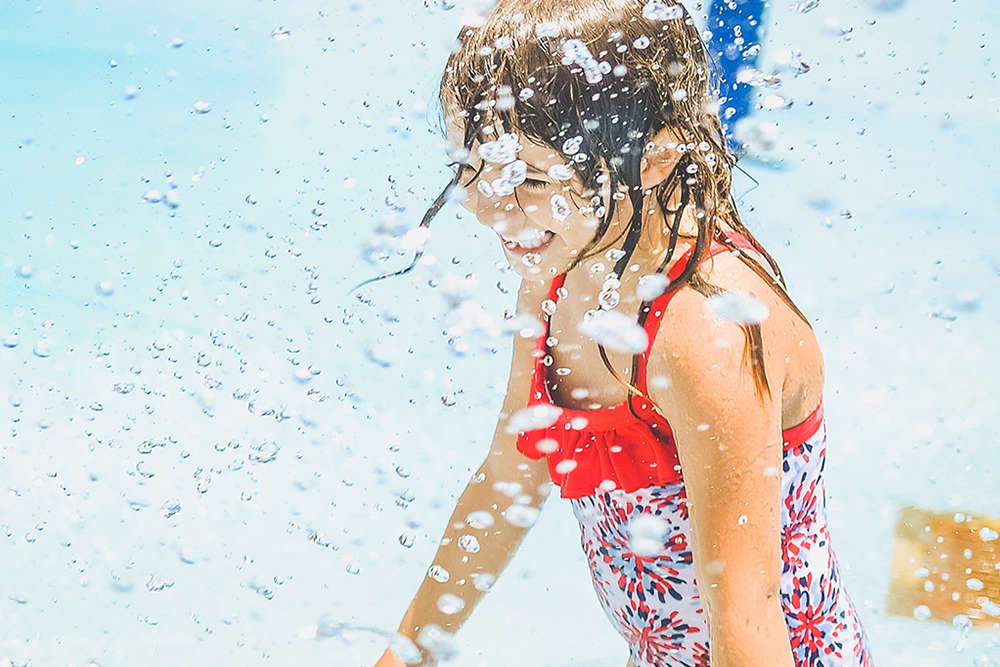 Young girl in a red and white swimsuit smiling and playing in water splashes.