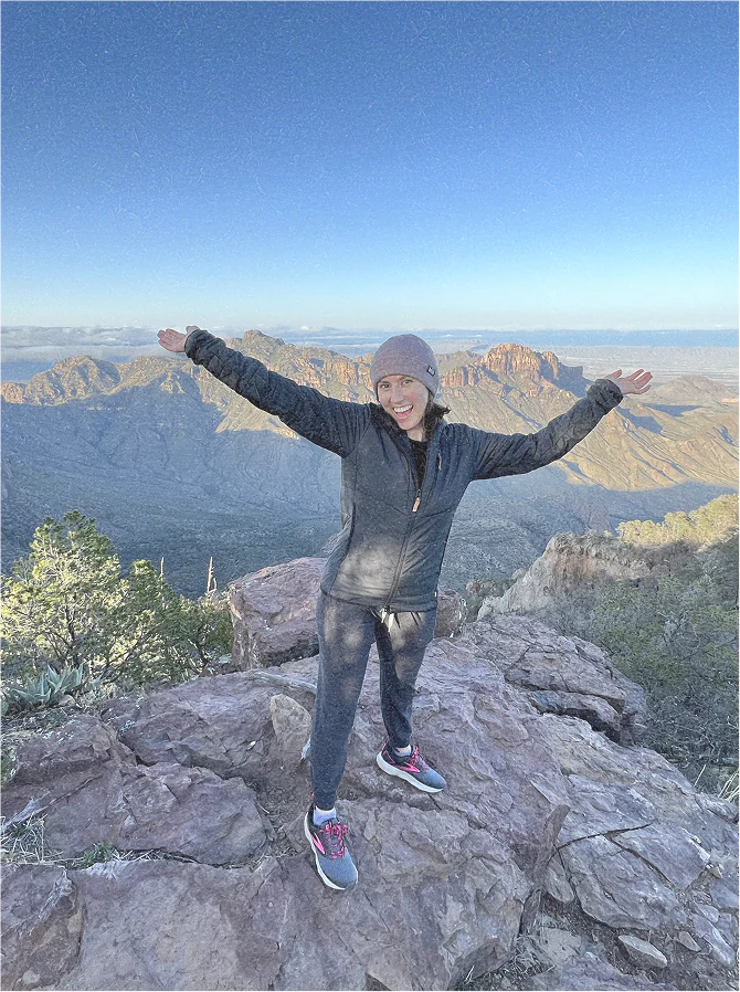 Donnine, one of our founders, posing on top of a mountain with her arms out.