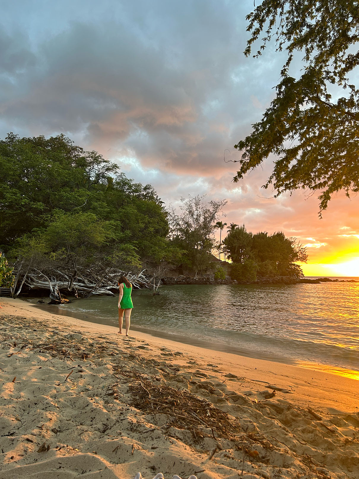 The sky glows with warm hues of orange and pink as the sun sets over the tranquil cove. A woman in a bright green dress stands barefoot at the shoreline, her gaze on the waves softly lapping at the sand. Driftwood and natural debris add texture to the beach, while the golden light of sunset bathes the scene in a serene, dreamlike glow.