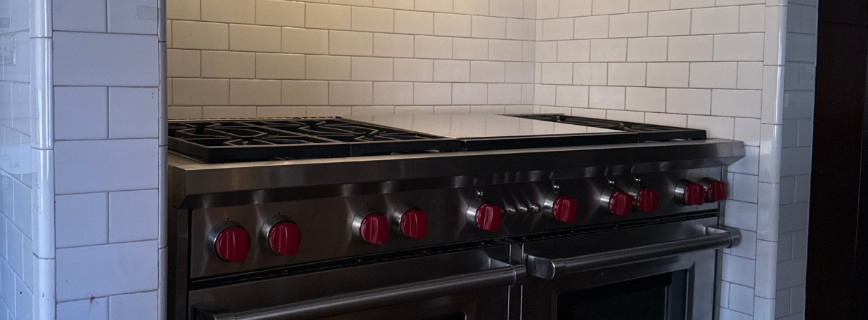 Large stainless steel commercial kitchen stove with red knobs, multiple burners, and double ovens, set against white tiled walls.