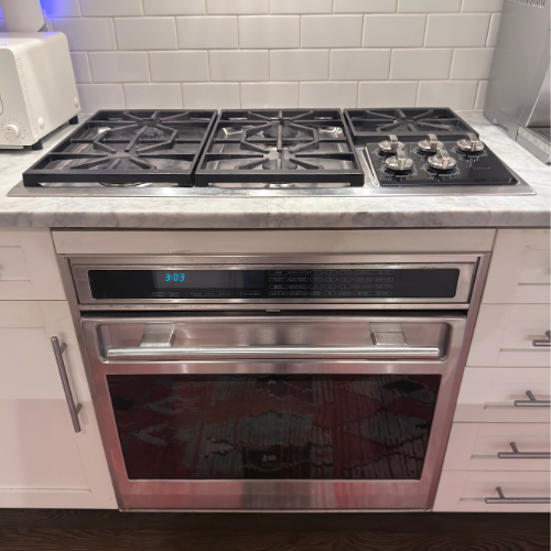 Built-in stainless steel oven with digital clock showing 3:03, and a gas stovetop with five burners and control knobs on a marble countertop.