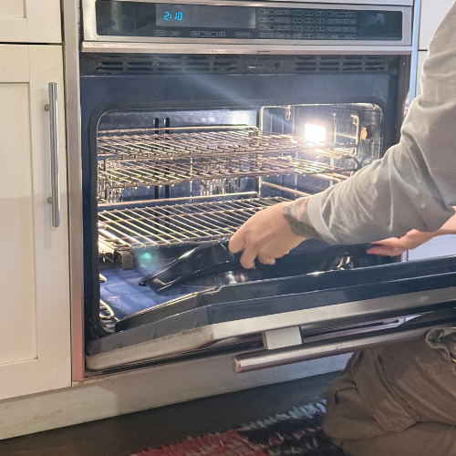 Stainless steel gas stove with four burners and a central griddle, featuring six control knobs and an oven beneath.