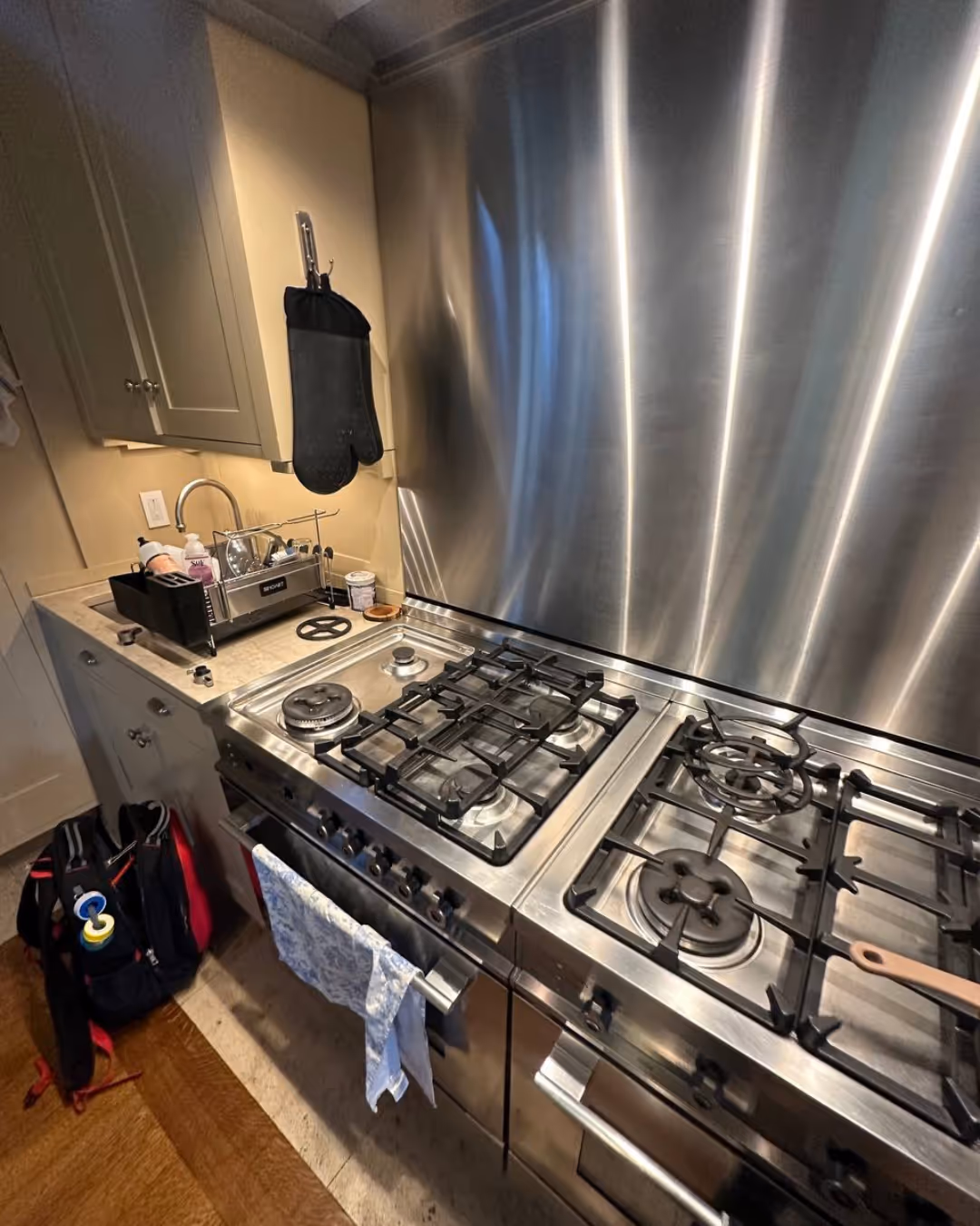 Man with a mustache wearing a blue jacket standing with arms crossed in a kitchen next to a stainless steel stove with red knobs.