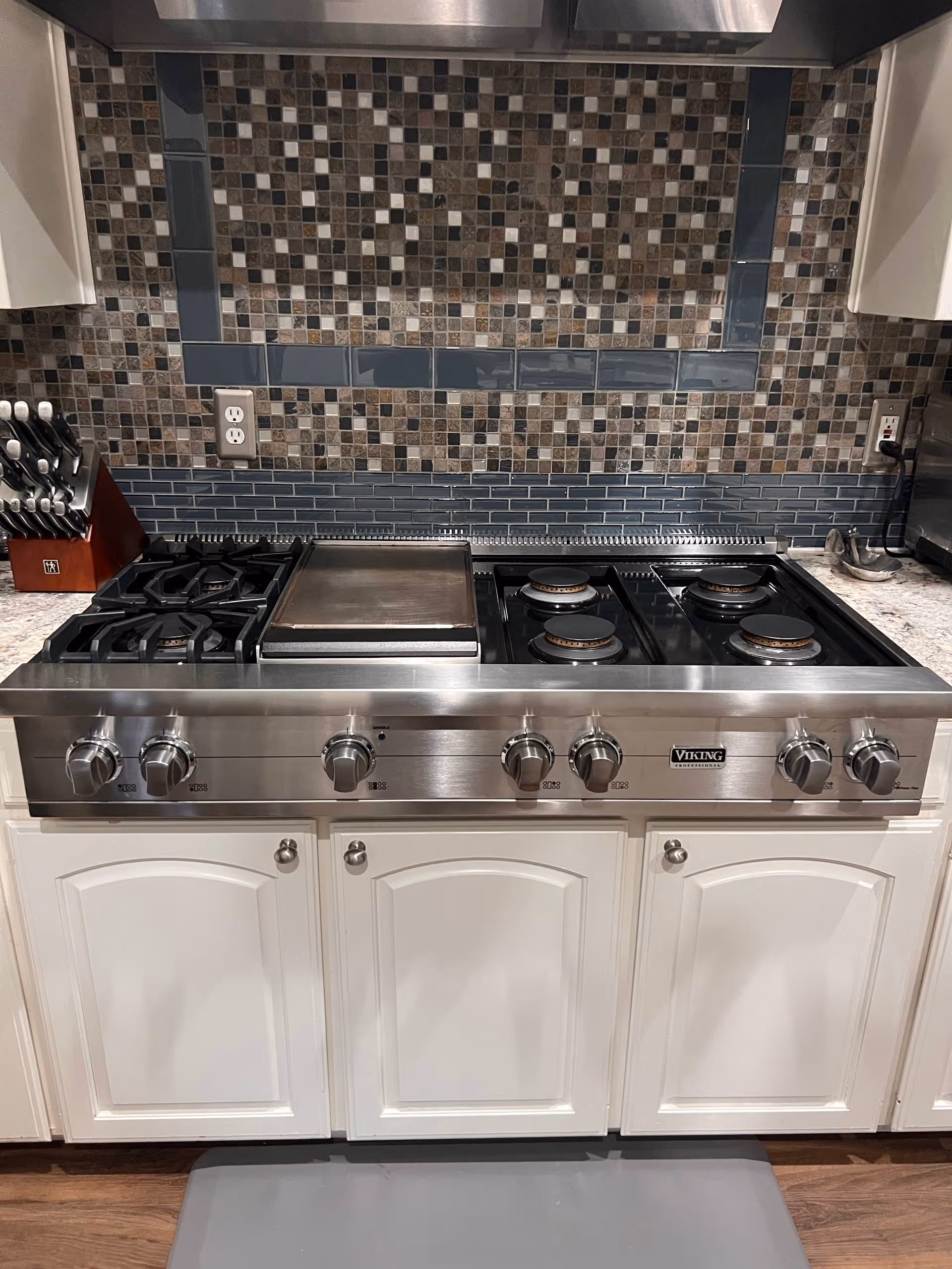 Stainless steel Viking Professional gas stove with six burners and griddle, set into white cabinetry with tiled backsplash.