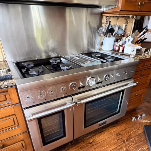Stainless steel gas stove with six burners and double oven doors in a kitchen with wooden cabinets and granite countertops.