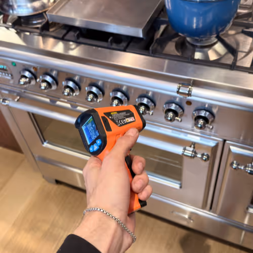 Hand holding an orange infrared thermometer pointed at a stainless steel gas stove with control knobs and a blue pot on a burner.