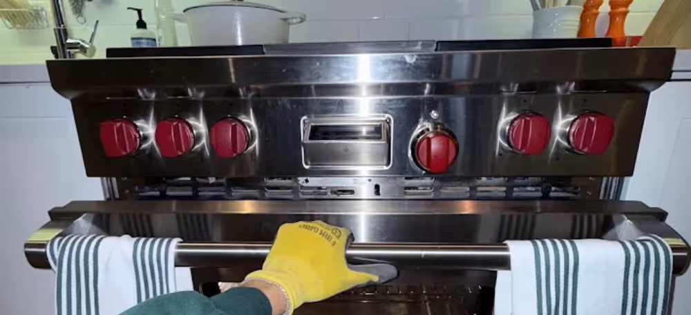 Person wearing a yellow glove opening a stainless steel oven with red control knobs and striped kitchen towels hanging on the handle.