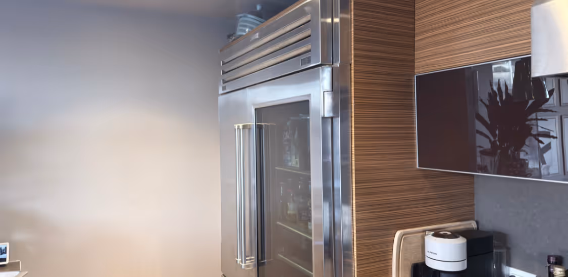 Stainless steel refrigerator with glass door in a modern kitchen with wood-paneled walls and a coffee maker on the counter.