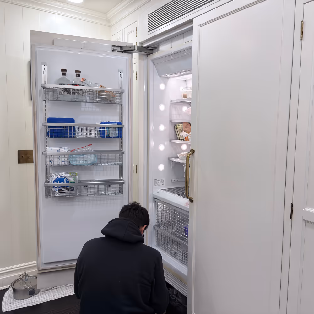 Technician repairing a built-in refrigerator with the door open in a kitchen.