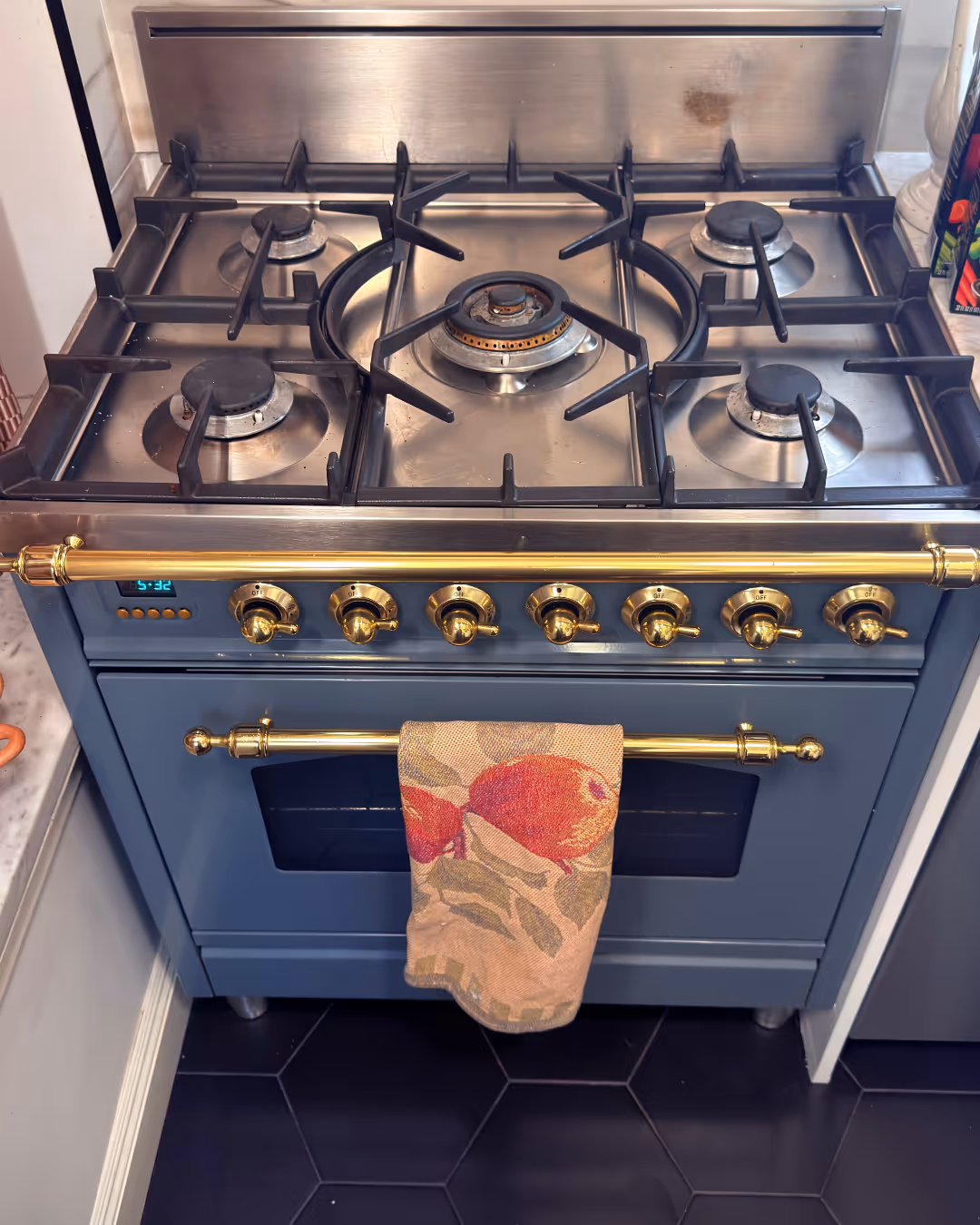 Blue gas stove with six burners, gold control knobs, and a floral-patterned towel hanging on the oven handle.