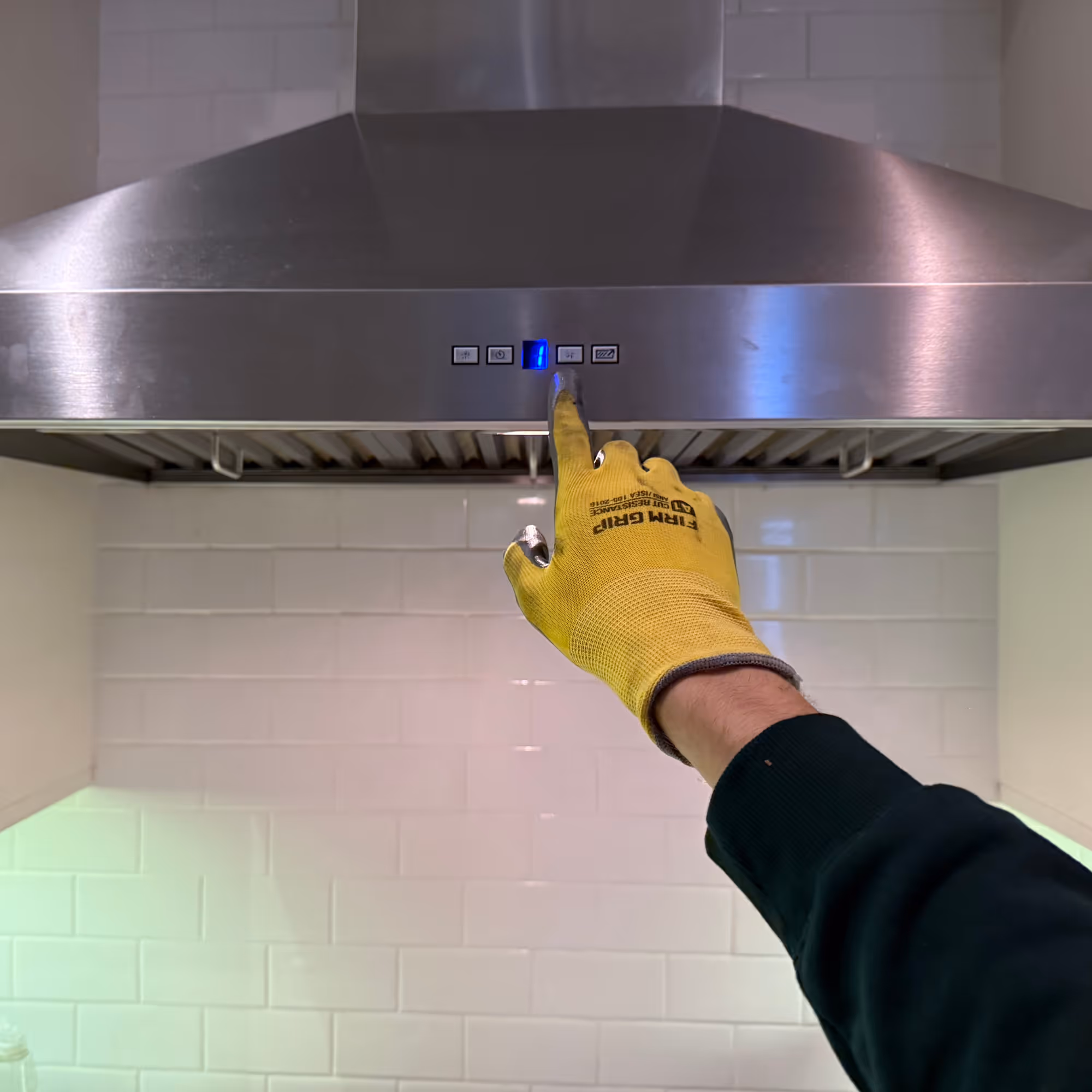 Person wearing a yellow work glove pressing a button on a stainless steel kitchen range hood control panel.